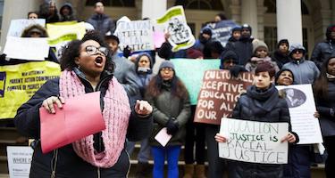 A Black woman wearing pink scarf is leading an educational justice rally for culturally responsive education in NYC Schools