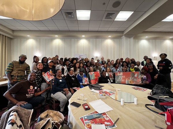 Large group photo of all the attendees of the Serve Our Sisters event. They are smiling and holding up a Serve Our Sisters event banner.