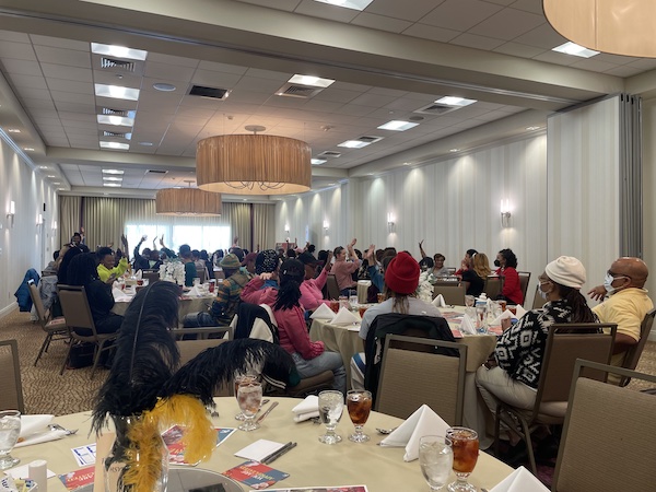 Attendees sit at tables in the event conference room. Hands are raised as they are participating in a game before enjoying a meal together. 