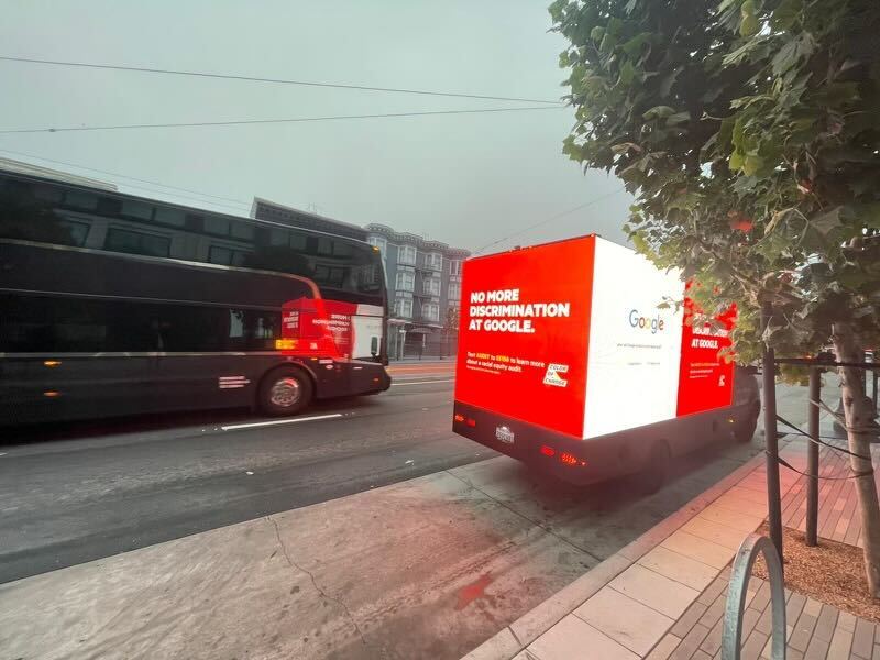 A photo of a mobile billboard near a Google campus shuttle. The mobile billboard says "No More Discrimination at Google."