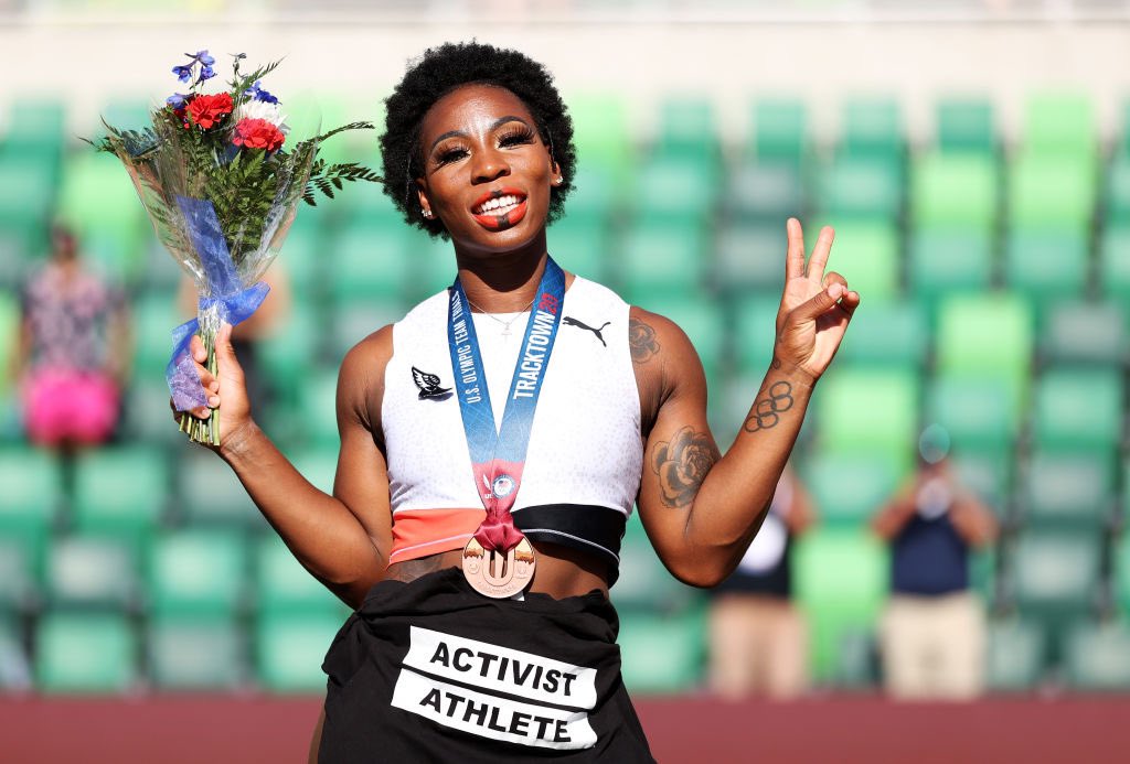 Gwen Berry at the U.S. Olympic Track and Field Trials. She is wearing her medal and has a shirt wrapped around her waist that says "Activist Athlete." She is holding flowers in one hand and doing a peace sign with her other hand. 